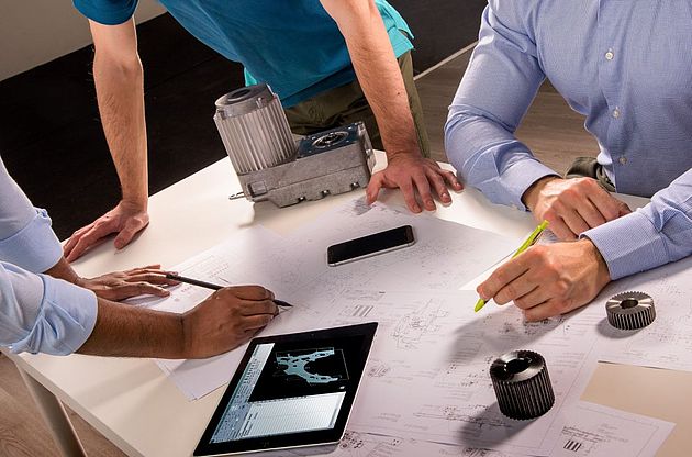 Three people standing at a table, machine parts and drawings in focus